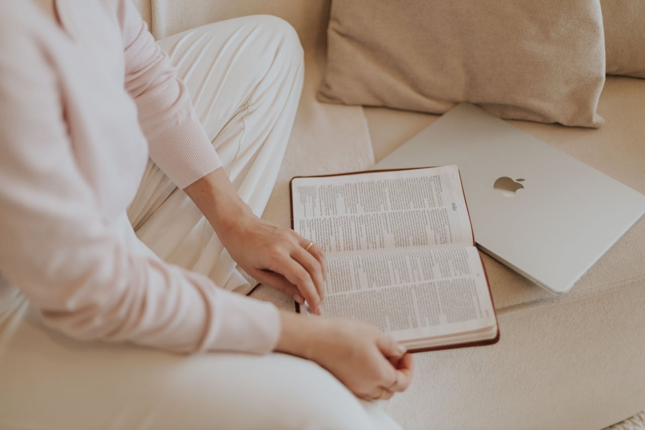 A woman in casual attire reads a book beside a laptop while sitting on a cozy sofa.